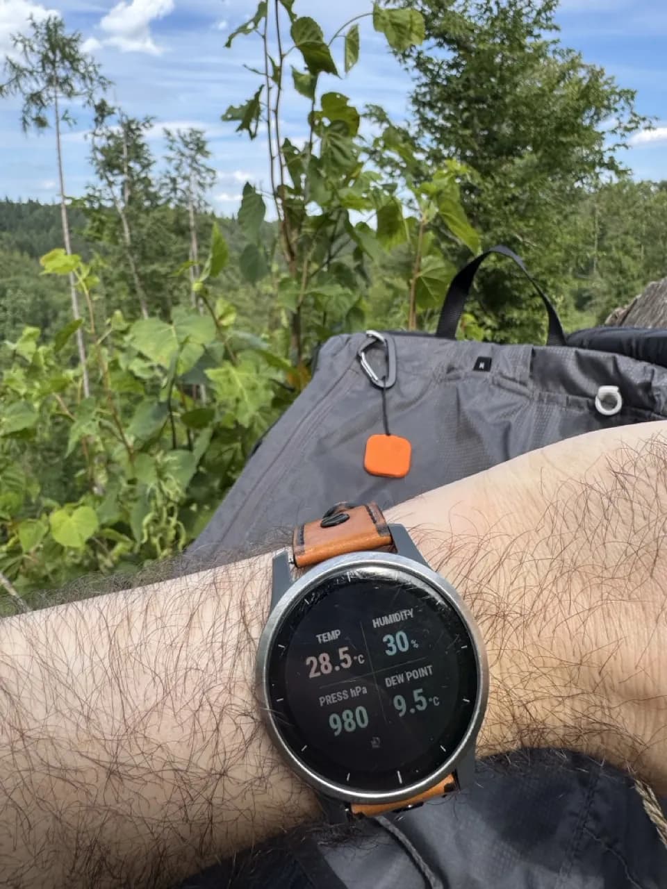 Thermio sensor clipped to a backpack while a Garmin watch shows live temperature, humidity, pressure, and dew point on the trail
